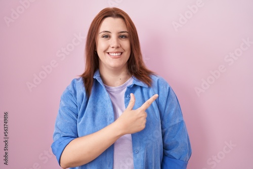 Young hispanic woman with red hair standing over pink background cheerful with a smile on face pointing with hand and finger up to the side with happy and natural expression