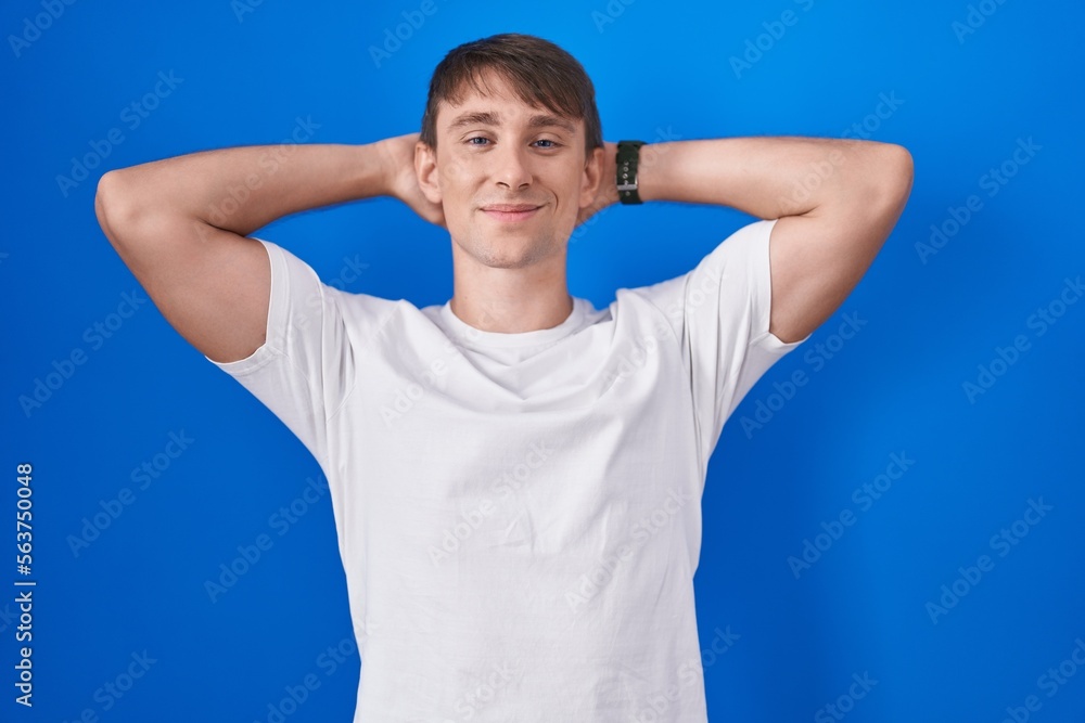 Caucasian blond man standing over blue background relaxing and stretching, arms and hands behind head and neck smiling happy Caucasian blond man standing over blue background relaxing and stretching, arms and hands behind head and neck smiling happy