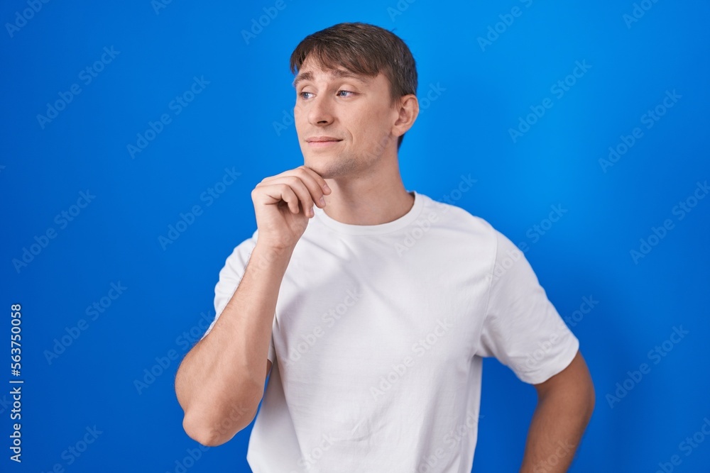 Caucasian blond man standing over blue background with hand on chin thinking about question, pensive expression. smiling with thoughtful face. doubt concept. Caucasian blond man standing over blue background with hand on chin thinking about question, pensive expression. smiling with thoughtful face. doubt concept.