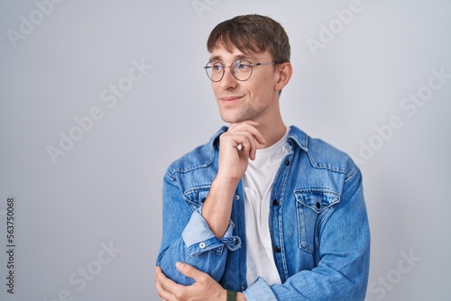 Caucasian blond man standing wearing glasses with hand on chin thinking about question, pensive expression. smiling and thoughtful face. doubt concept.