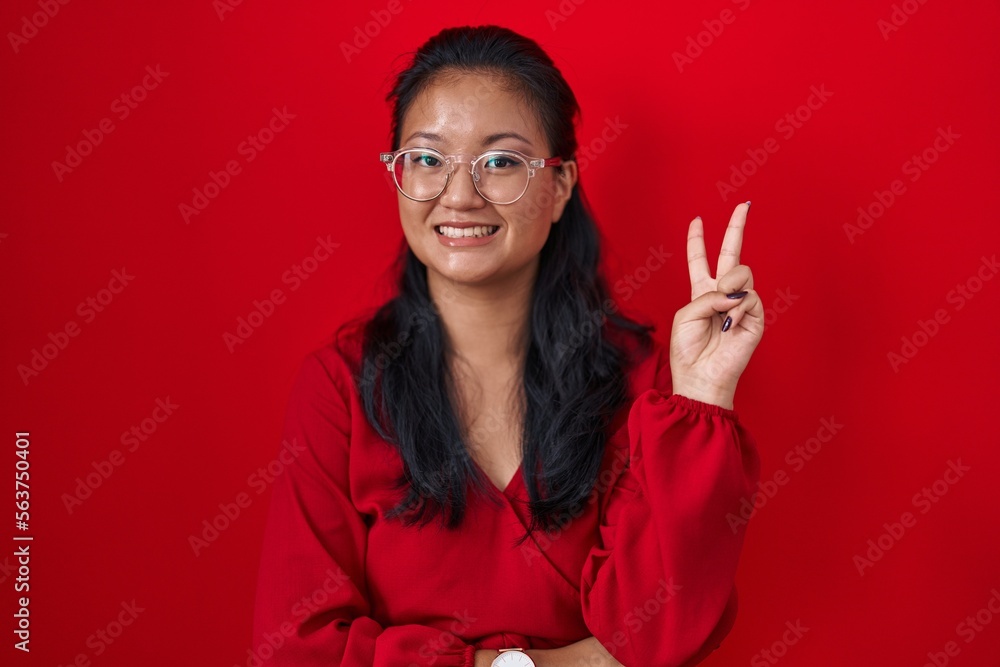 Asian young woman standing over red background smiling with happy face winking at the camera doing victory sign with fingers. number two. Asian young woman standing over red background smiling with happy face winking at the camera doing victory sign with fingers. number two.