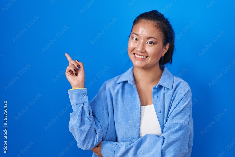 Asian young woman standing over blue background with a big smile on face, pointing with hand finger to the side looking at the camera. Asian young woman standing over blue background with a big smile on face, pointing with hand finger to the side looking at the camera.