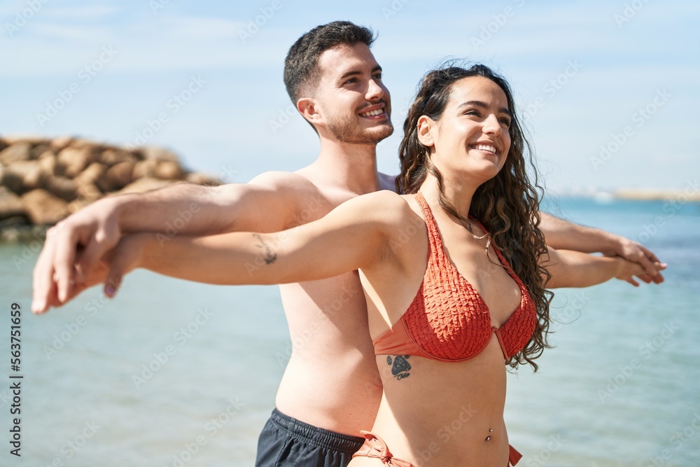 Young hispanic couple tourists wearing swimsuit standing with arms open at seaside