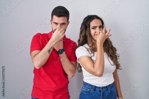 Young hispanic couple standing over isolated background smelling something stinky and disgusting, intolerable smell, holding breath with fingers on nose. bad smell