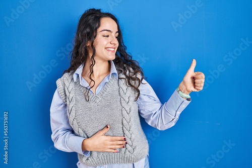 Young brunette woman standing over blue background looking proud, smiling doing thumbs up gesture to the side