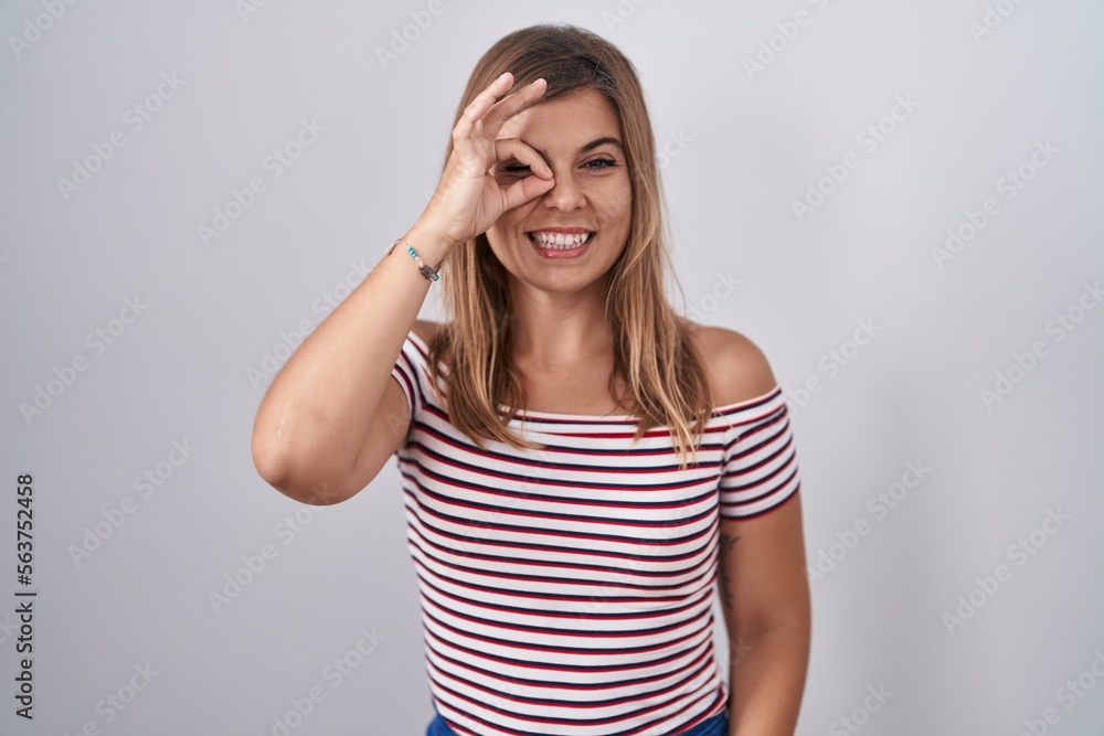 Young hispanic woman standing over isolated background doing ok gesture with hand smiling, eye looking through fingers with happy face. Young hispanic woman standing over isolated background doing ok gesture with hand smiling, eye looking through fingers with happy face.