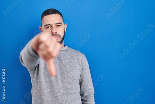 Hispanic man standing over blue background looking unhappy and angry showing rejection and negative with thumbs down gesture. bad expression.