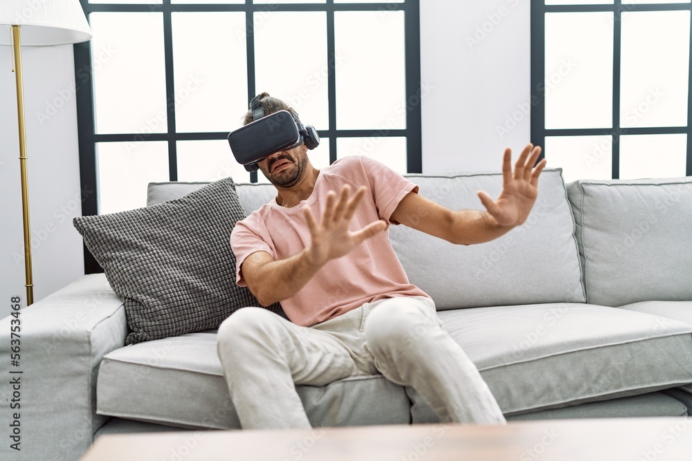Fototapeta premium Handsome hispanic man afraid wearing virtual reality glasses at the living room at home
