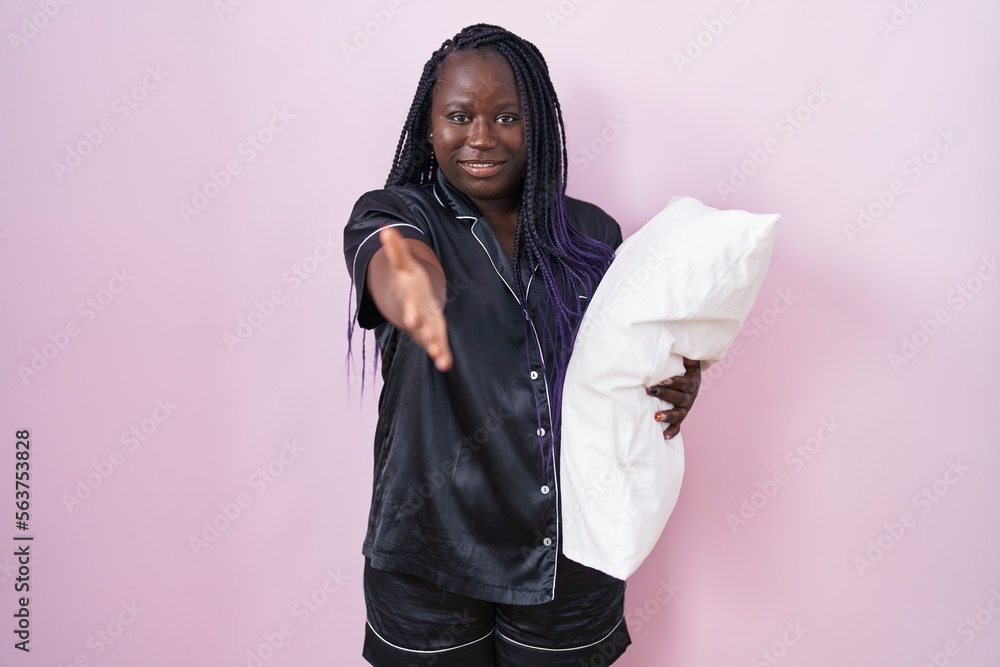 Young african woman wearing pijama hugging pillow smiling cheerful offering palm hand giving assistance and acceptance. Young african woman wearing pijama hugging pillow smiling cheerful offering palm hand giving assistance and acceptance.