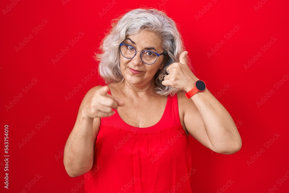 Middle age woman with grey hair standing over red background smiling doing talking on the telephone gesture and pointing to you. call me. Middle age woman with grey hair standing over red background smiling doing talking on the telephone gesture and pointing to you. call me.