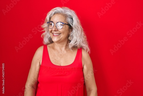 Middle age woman with grey hair standing over red background looking away to side with smile on face, natural expression. laughing confident.