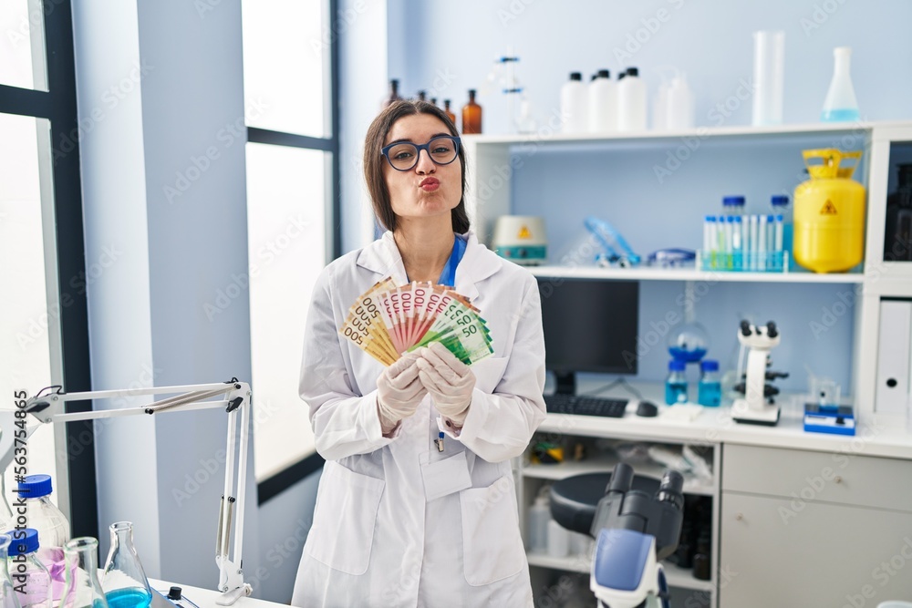 Young hispanic woman working at scientist laboratory holding money banknotes looking at the camera blowing a kiss being lovely and sexy. love expression. Young hispanic woman working at scientist laboratory holding money banknotes looking at the camera blowing a kiss being lovely and sexy. love expression.