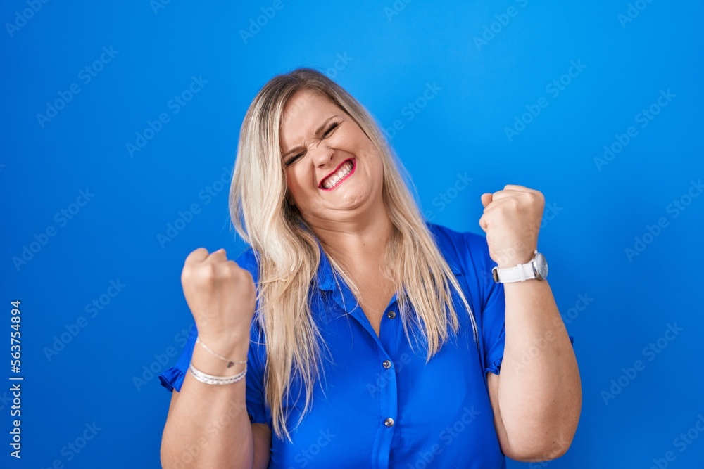 Fototapeta premium Caucasian plus size woman standing over blue background very happy and excited doing winner gesture with arms raised, smiling and screaming for success. celebration concept.