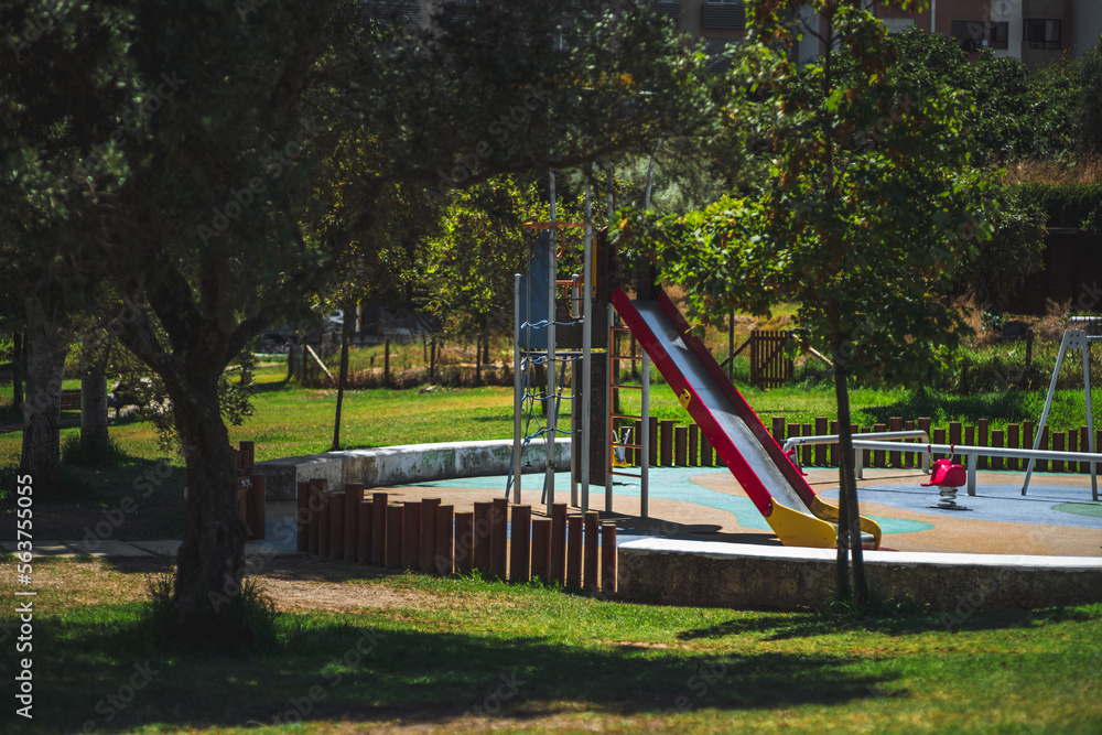Fototapeta premium A shot of a children's playground, focusing on a colorful slide, besides some of the swings, this in a round area partially enclosed by wood and cement, bordered by a lawn with shady trees
