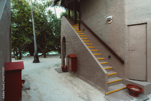 Tropical terracotta colored house villa housing entrance with sandy flooring  a red firefighting tool  and bowl for foot washing  yellow stripes highlighting the steps of the staircase