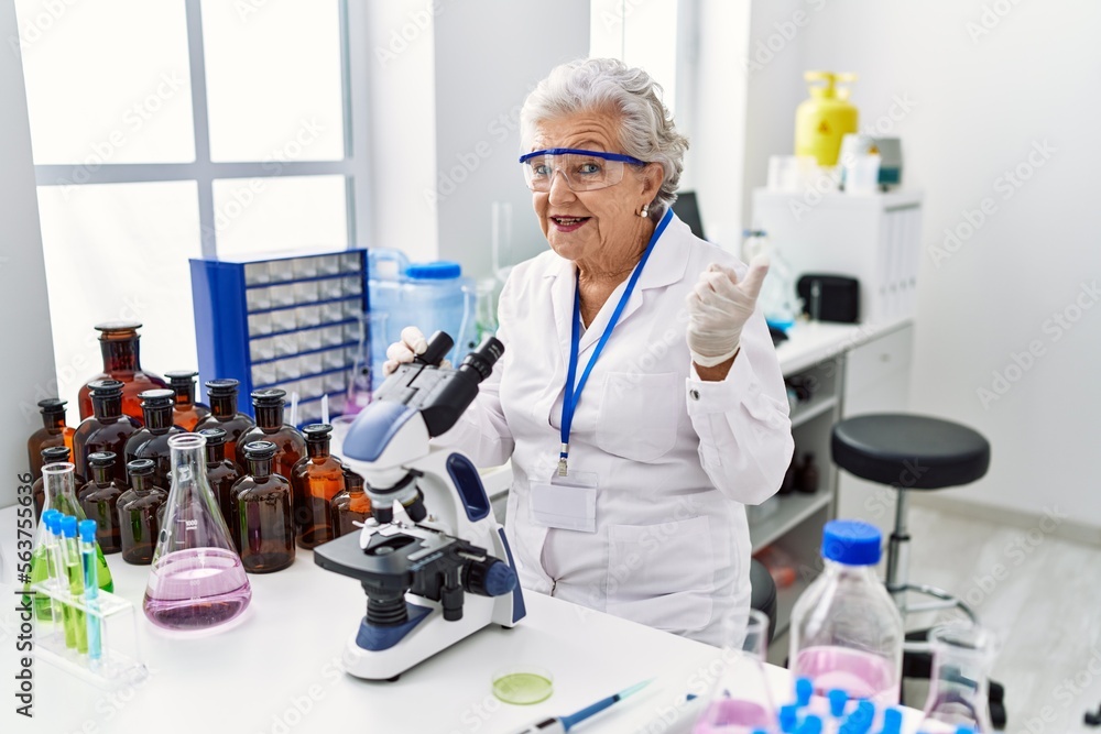 Senior woman with grey hair working at scientist laboratory smiling happy pointing with hand and finger to the side Senior woman with grey hair working at scientist laboratory smiling happy pointing with hand and finger to the side