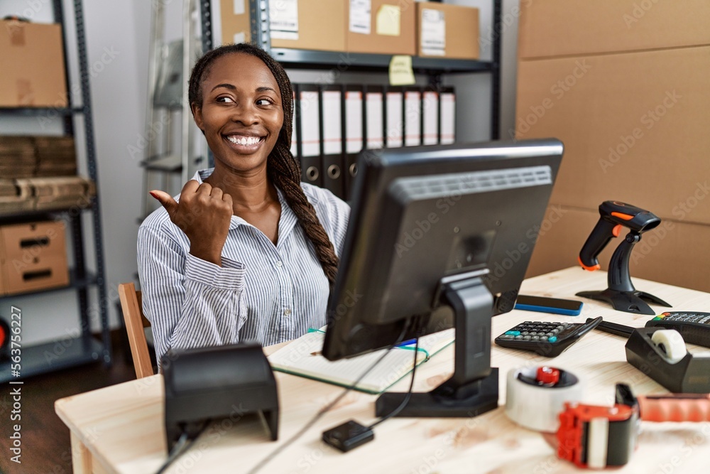 African woman working at small business ecommerce smiling with happy face looking and pointing to the side with thumb up. African woman working at small business ecommerce smiling with happy face looking and pointing to the side with thumb up.