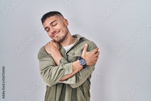 Foto Young hispanic man standing over isolated background hugging oneself happy and positive, smiling confident