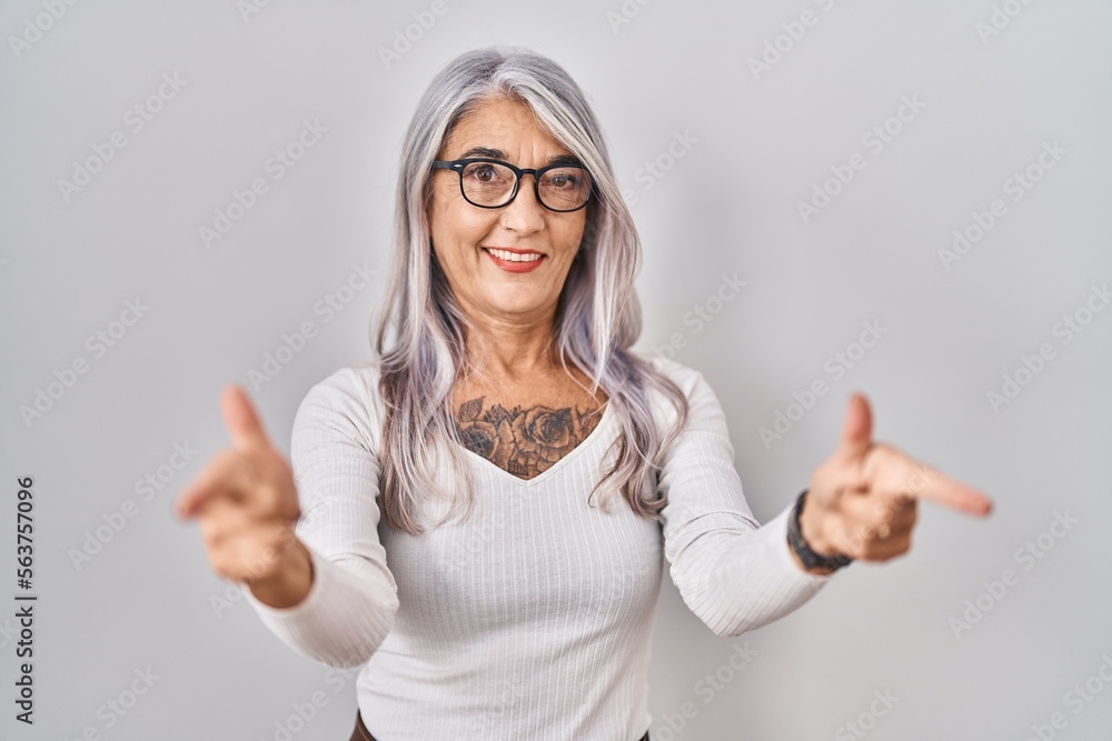 Middle age woman with grey hair standing over white background pointing fingers to camera with happy and funny face. good energy and vibes. Middle age woman with grey hair standing over white background pointing fingers to camera with happy and funny face. good energy and vibes.