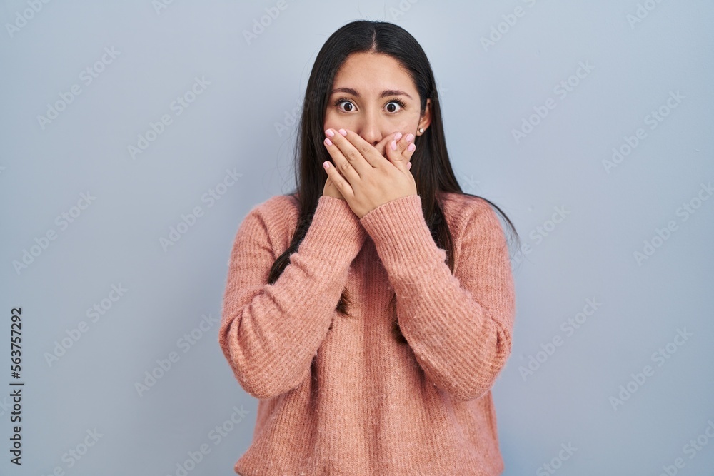 Young latin woman standing over blue background shocked covering mouth with hands for mistake. secret concept. Young latin woman standing over blue background shocked covering mouth with hands for mistake. secret concept.