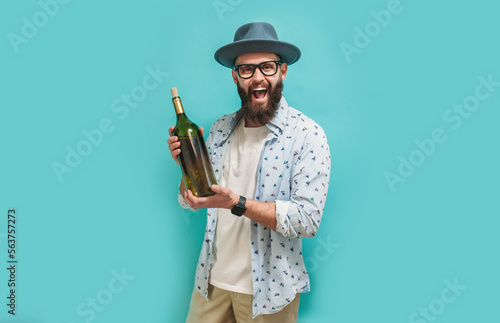 Young bearded charismatic hipster somilier showing a big bottle of wine, isolated on a blue studio background. Wine presentation. photo