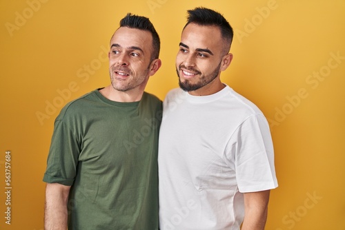Homosexual couple standing over yellow background looking away to side with smile on face, natural expression. laughing confident.