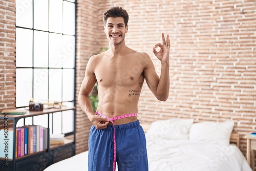 Young hispanic man using tape measure measuring waist doing ok sign with fingers, smiling friendly gesturing excellent symbol