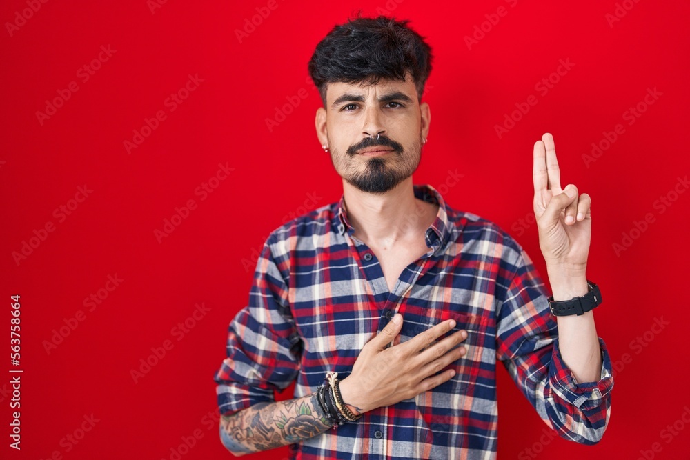 Fototapeta premium Young hispanic man with beard standing over red background smiling swearing with hand on chest and fingers up, making a loyalty promise oath