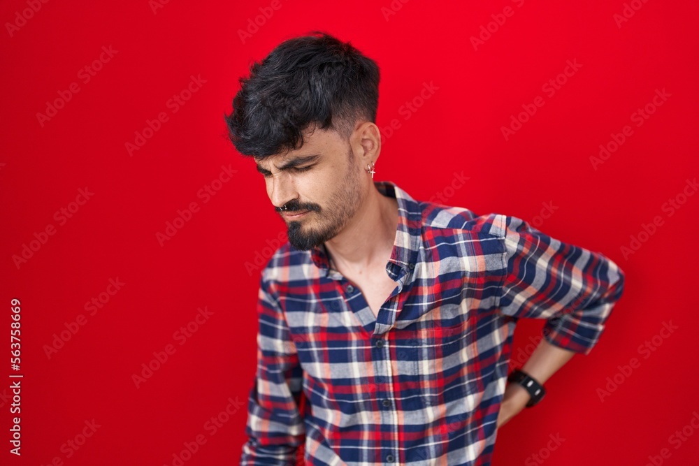 Young hispanic man with beard standing over red background suffering of backache, touching back with hand, muscular pain Young hispanic man with beard standing over red background suffering of backache, touching back with hand, muscular pain