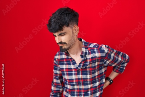 Young hispanic man with beard standing over red background suffering of backache, touching back with hand, muscular pain