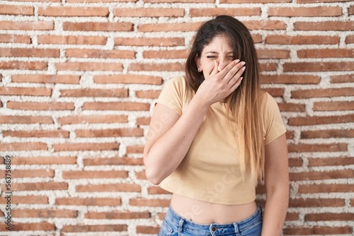 Young brunette woman standing over bricks wall smelling something stinky and disgusting, intolerable smell, holding breath with fingers on nose. bad smell