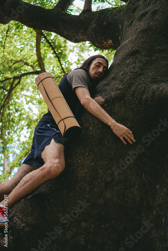 Young latin hippie man hugging a tree in a wooded area with a yoga mat hanging. Vertical photo.