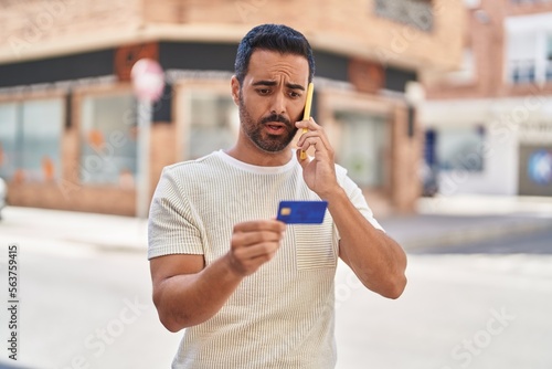 Young hispanic man talking on the smartphone and using credit card at street