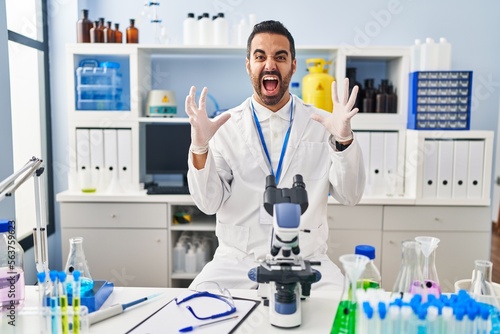Young hispanic man with beard working at scientist laboratory crazy and mad shouting and yelling with aggressive expression and arms raised. frustration concept.