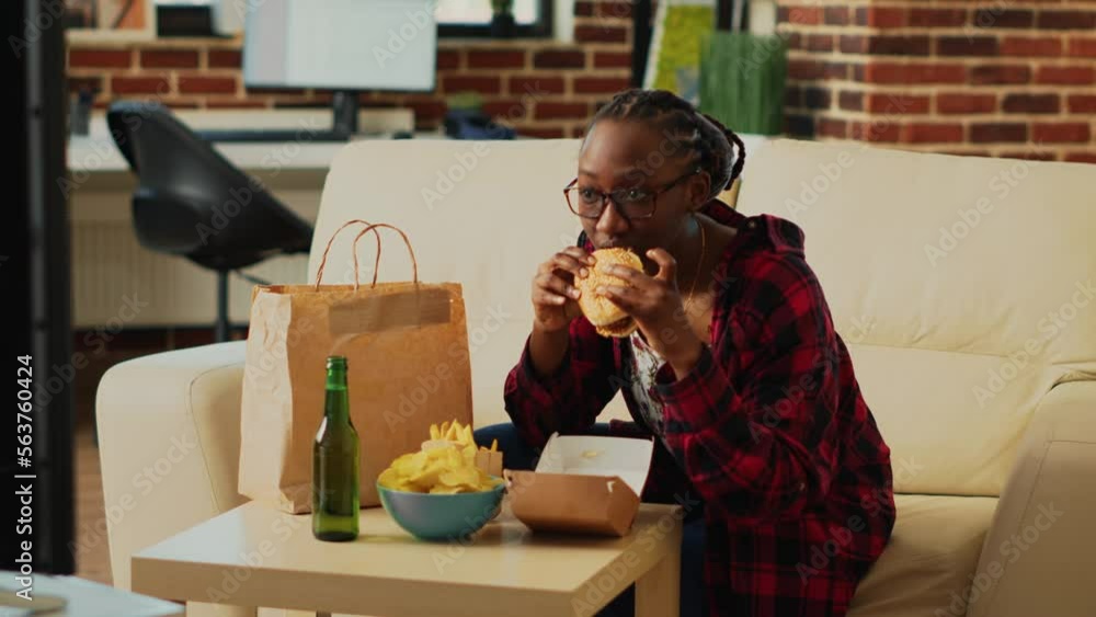 Happy woman eating hamburgers with fries and beer, having fun with action film on television. Smiling person enjoying burger and snacks in bowl, binge watching tv show in living room. Happy woman eating hamburgers with fries and beer, having fun with action film on television. Smiling person enjoying burger and snacks in bowl, binge watching tv show in living room.