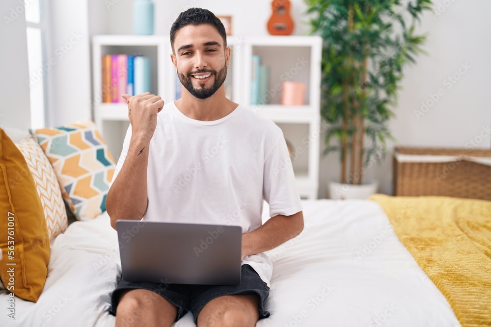 Hispanic young man using computer laptop sitting on the bed pointing thumb up to the side smiling happy with open mouth Hispanic young man using computer laptop sitting on the bed pointing thumb up to the side smiling happy with open mouth