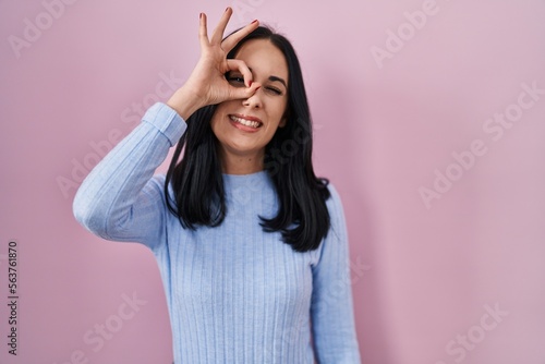 Hispanic woman standing over pink background doing ok gesture with hand smiling, eye looking through fingers with happy face.