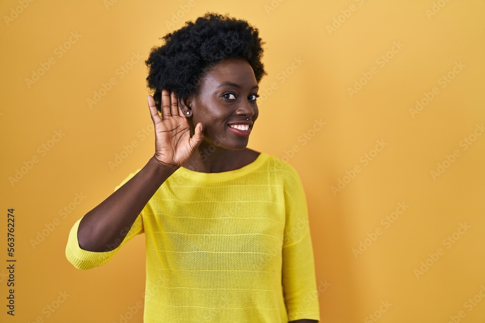 African young woman standing over yellow studio smiling with hand over ear listening an hearing to rumor or gossip. deafness concept. African young woman standing over yellow studio smiling with hand over ear listening an hearing to rumor or gossip. deafness concept.