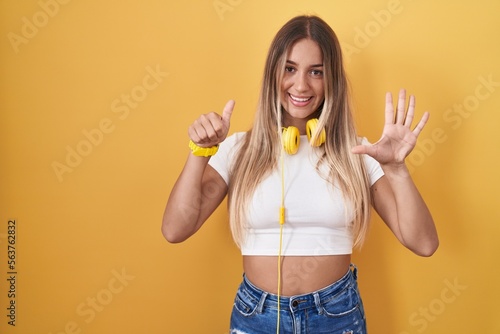 Young blonde woman standing over yellow background wearing headphones showing and pointing up with fingers number six while smiling confident and happy.