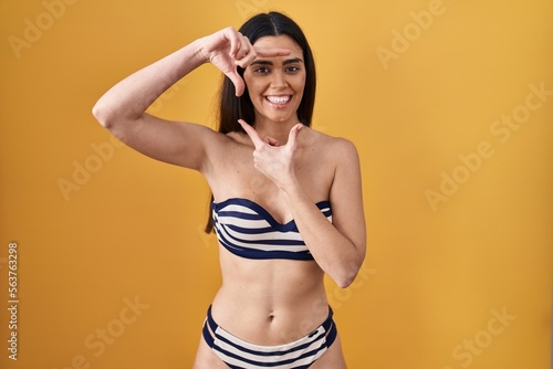 Young brunette woman wearing bikini over yellow background smiling making frame with hands and fingers with happy face. creativity and photography concept.