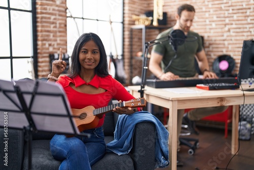 Young indian woman playing ukulele at music studio surprised with an idea or question pointing finger with happy face, number one