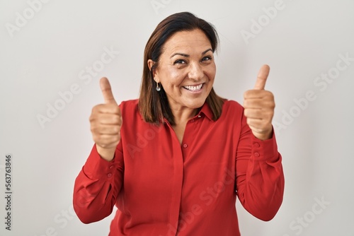 Hispanic mature woman standing over white background success sign doing positive gesture with hand, thumbs up smiling and happy. cheerful expression and winner gesture.