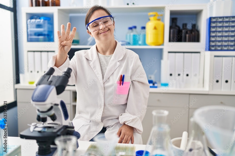 Hispanic girl with down syndrome working at scientist laboratory showing and pointing up with fingers number four while smiling confident and happy.