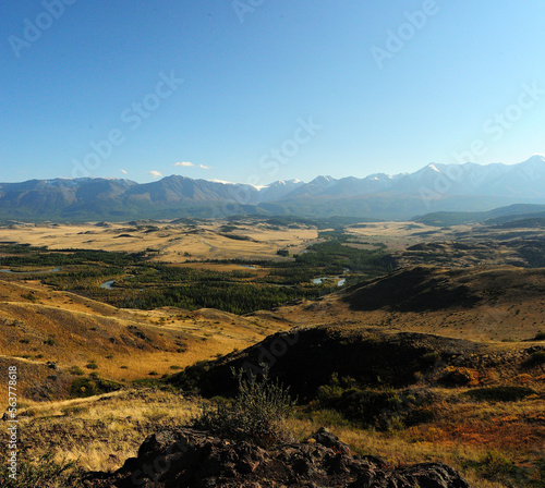 View from a high hill to the winding bed of a beautiful river with a dense coniferous forest in a picturesque steppe at the foot of a mountain range with snow-capped peaks.