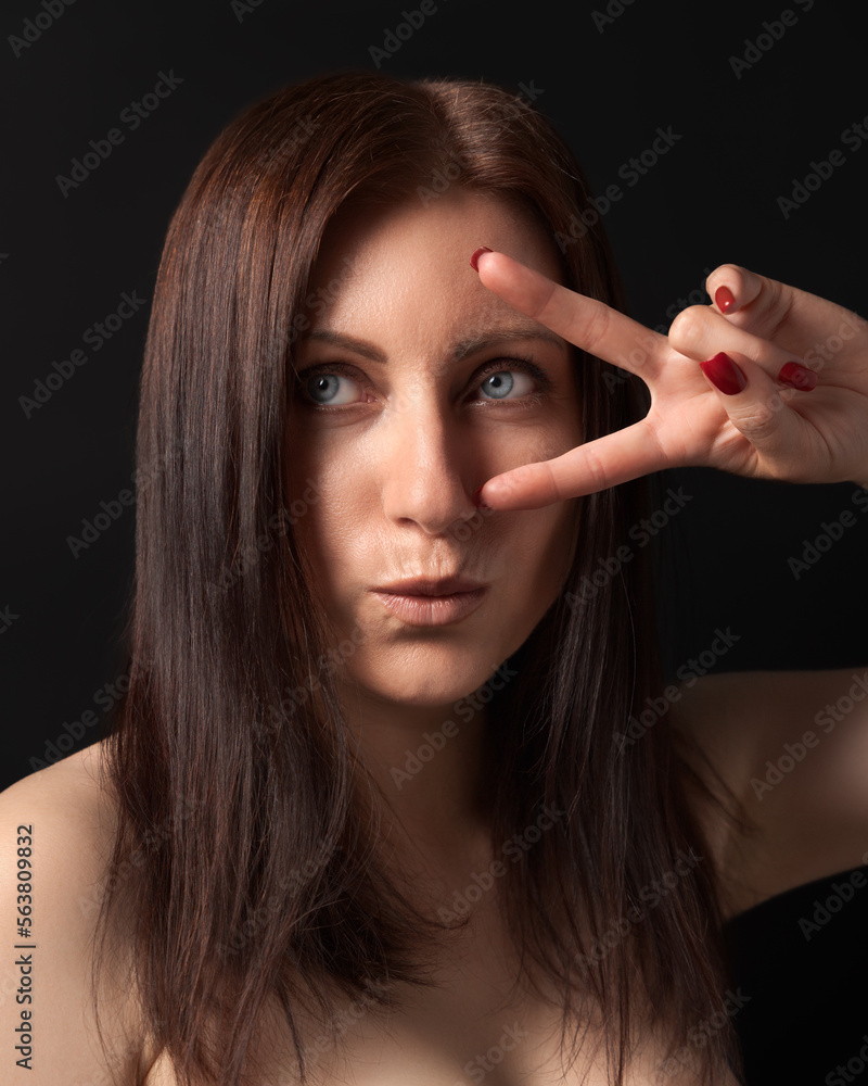 Obraz premium Studio portrait of 40-year-old woman showing victory or peace gesture with fingers near gray eyes. Caucasian ethnicity brunette with long hair and outstretched lips looking away on black background.