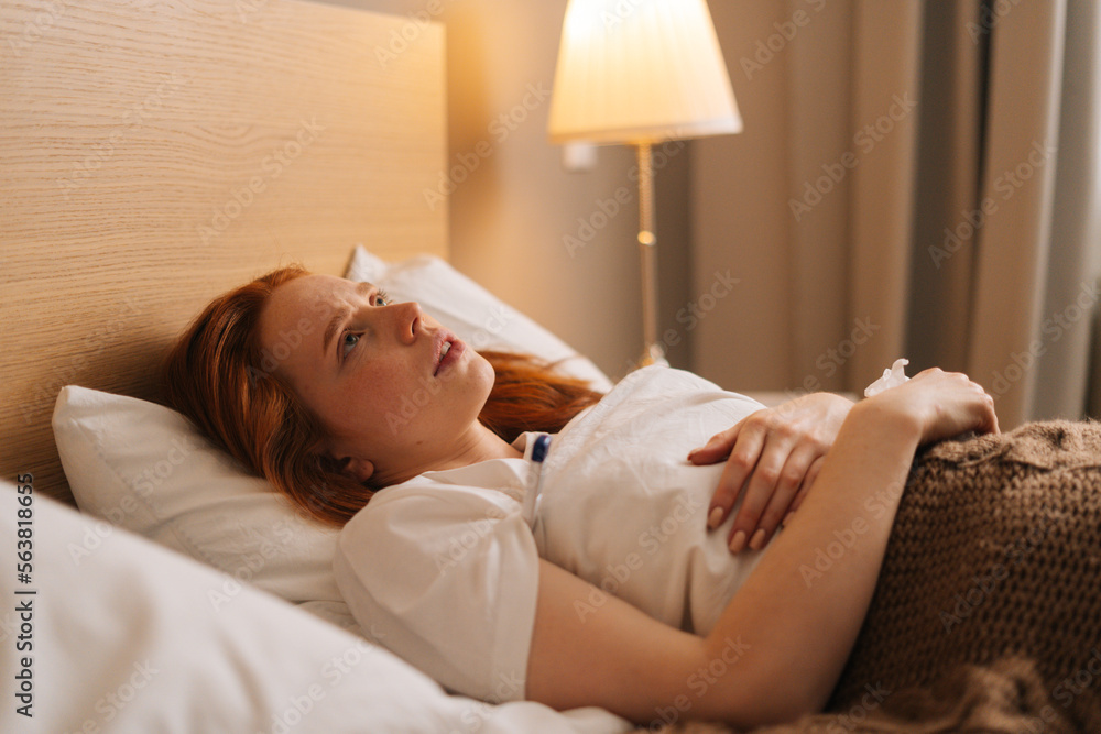 Side view of sick exhausted young woman with flu lying under blanket on