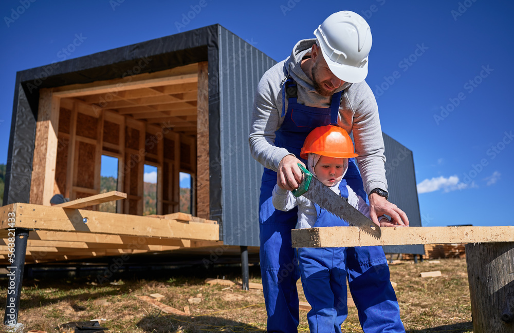 Father with toddler son building wooden frame house in the Scandinavian style barnhouse. Male builders using hand saw to cut boards on construction site on sunny day. Carpentry and family concept. Father with toddler son building wooden frame house in the Scandinavian style barnhouse. Male builders using hand saw to cut boards on construction site on sunny day. Carpentry and family concept.