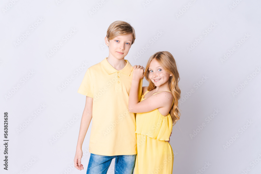 Two happy children standing near an empty gray background and hugging cute little girl with long hair is hugging cute blonde girl showing her love and care Brother and sister have fun posing in studio Two happy children standing near an empty gray background and hugging cute little girl with long hair is hugging cute blonde girl showing her love and care Brother and sister have fun posing in studio
