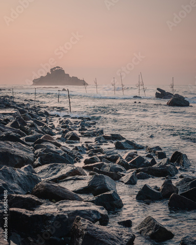 Ahangama, Sri Lanka : view from the beach with rocks in the foreground and an islet in the background at sunrise. Traditional fishermen sticks in the water. photo