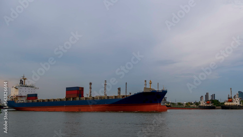 transportation logistics cargo port carrier almost. empty red and blue containership transporting its goods over the river with the harbour in the background. The bulbous bow and prow in red